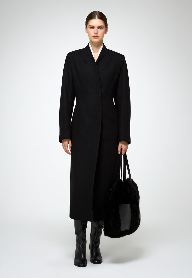 Cappotto lungo nero con taglio dritto, colletto alto e chiusura frontale. Abbinato a stivali neri al ginocchio e una morbida borsa nera.