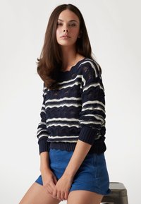 Maglione a righe blu navy e bianche in maglia con un motivo ondulato, polsini a coste e scollo ampio, abbinato a pantaloni corti blu.
