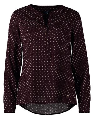 Femme marchant portant un chemisier noir à manches longues avec des pois blancs, un pantalon noir et des chaussures à talons noirs devant un fond uni.