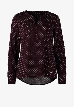 Kvinde går iført en sort bluse med lange ærmer med hvide prikker, sorte bukser og sorte højhælede sko mod en ensfarvet baggrund.