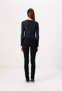 Kvinna med långt rakt hår står vänd mot en vit vägg, iförd en svart långärmad topp, svarta jeans och mörka sneakers med beige sulor.