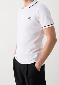 Valkoinen polo-paita, jossa on mustat reunukset kauluksessa ja hihoissa. Paitassa on pieni musta "CK" -logo vasemmalla rinnalla. Puuvillakangasta, lyhyet hihat.