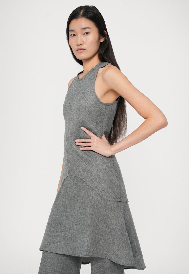 Femme avec de longs cheveux noirs portant une robe grise asymétrique sans manches par-dessus un pantalon assorti, posant avec une main sur la hanche devant un fond blanc.