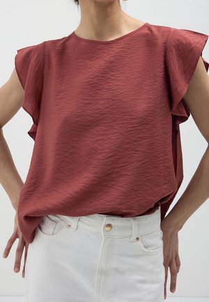 Frau trägt eine rostfarbene, strukturierte ärmellose Bluse mit Rüschen an den Schultern, in weiße Jeans mit goldenen Knöpfen gesteckt.