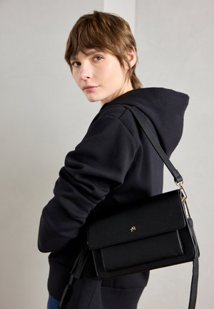 Junge Person mit kurzen braunen Haaren, die einen schwarzen Hoodie trägt und eine schwarze Umhängetasche mit goldener Schnalle trägt.