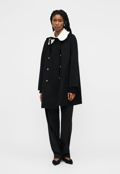 Γυναίκα με πλεξούδες που φοράει μαύρο διπλόπατο παλτό με λευκό γιακά, μαύρο παντελόνι και μαύρα μυτερά παπούτσια, στέκεται σε λευκό φόντο.