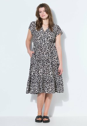 Frau steht, trägt ein knielanges, kurzärmeliges Kleid mit Leopardenmuster und schwarze offene Sandalen vor einem einfarbigen hellen Hintergrund.