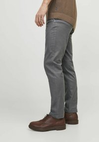 Hombre vestido con pantalones gris ajustados, zapatos de cuero marrón con cordones y un suéter marrón, de pie de lado sobre un fondo blanco.