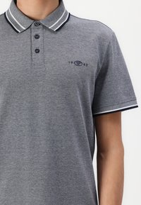 Grå polo t-shirt lavet af struktureret stof, med en krave med marineblå og hvide striber, tre knaplukninger og et lille logo på brystet.
