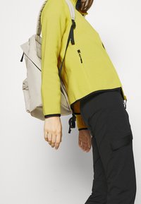 Gelbe Fleecejacke mit schwarzem Saum, mit Reißverschlusstasche, getragen mit schwarzen Hosen. Hellbeige Rucksack mit strukturiertem Stoff und Riemen.