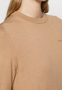 Närbild av en beige stickad tröja med runt halsringning och en liten broderad "BOSS"-logotyp på bröstet, som bärs av en person.