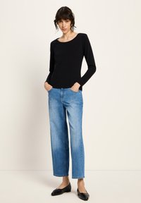 Eine Frau mit kurzen, dunklen Haaren, die ein schwarzes Langarmoberteil, hellblaue weit geschnittene Jeans und schwarze flache Schuhe trägt, steht mit den Händen in den Taschen.