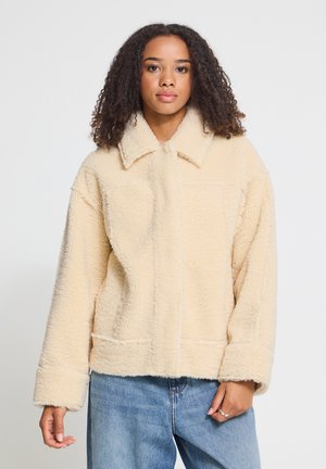 Jeune femme aux cheveux bouclés portant une veste crème duveteuse et un jean bleu, debout devant un fond clair uni.