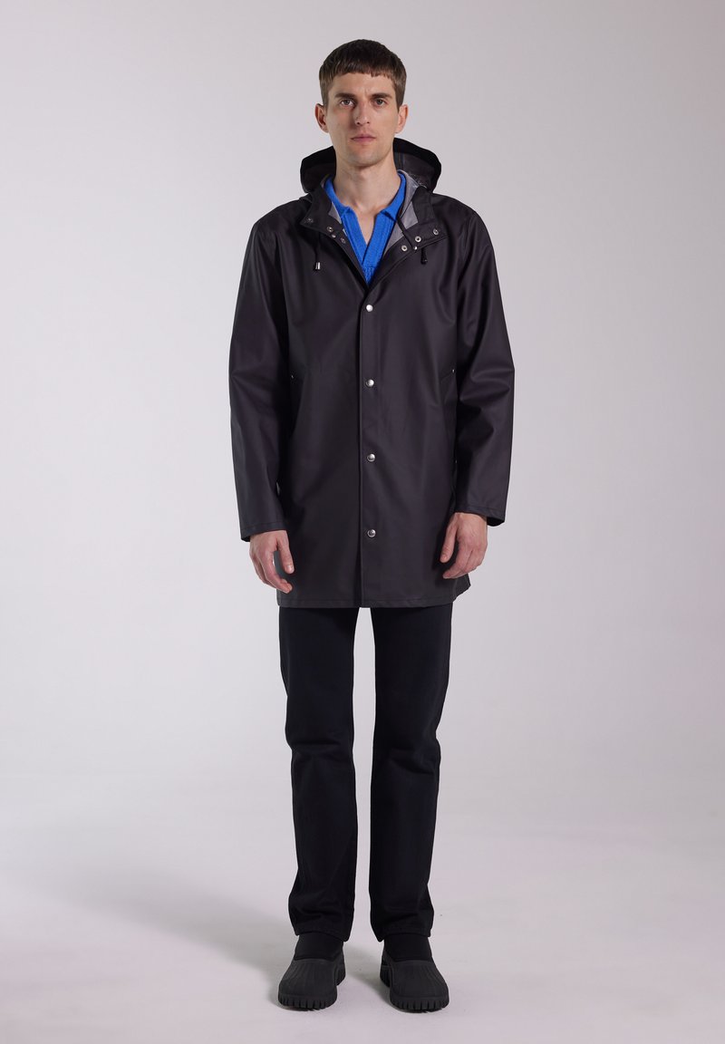 Black waterproof rain jacket with hood, snap buttons, and two side pockets; paired with blue shirt and black pants. Rubber shoes complete the outfit.