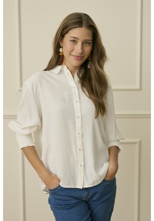 Mlada žena s dugom valovitom kosom, odjevena u široku bijelu košulju s dugmićima i plave traperice, s rukama u džepovima, stoji uz neutralni zid s panelima.