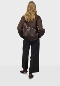 Braune Leder-Crossbody-Tasche mit glatter Textur und geschwungenem Design, getragen über einem braunen oversized Pullover und schwarzen weitgeschnittenen Hosen.