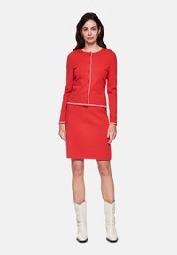 Red knitted two-piece set featuring a buttoned cardigan and a fitted skirt, accented with white trim. Model wears white ankle boots.