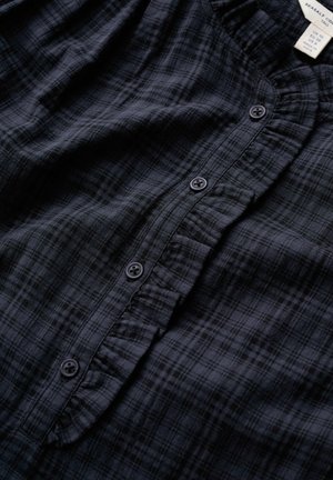 Tumši zila pleda krekls ar nelielu volānu ap pogu joslu un četrām redzamām pogām, kakla izgriezumā redzama izmēra un zīmola etiķete.