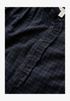 Tumši zila pleda krekls ar nelielu volānu ap pogu joslu un četrām redzamām pogām, kakla izgriezumā redzama izmēra un zīmola etiķete.