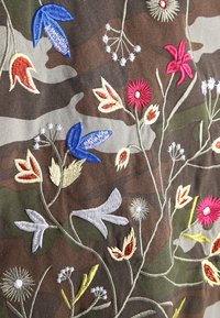 Broderad tyg med ett kamouflagemönster, dekorerad med färgglada blommönster i blå, röd, gul och vit tråd, på en strukturerad bakgrund.