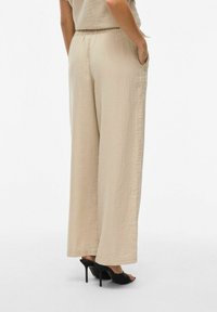 Pantalons larges beige en tissu léger, dotés d'une taille élastique, de poches latérales, et associés à des sandales noires à talons.