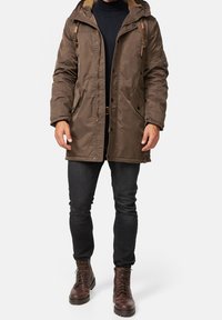 Bruine geïsoleerde jas met een met bont gevoerde capuchon, meerdere zakken en knoopsluitingen. Genomineerd over een zwarte trui en gecombineerd met donkere jeans en bruine laarzen.