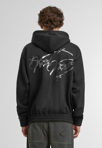 Schwarzer Hoodie mit einem silbernen Grafikdesign und Sternen auf dem Rücken, mit lockerer Passform und geripptem Bündchen. Weicher Stoff. 