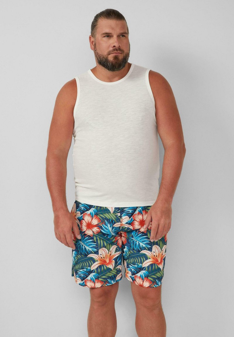 Witte mouwloze tanktop gecombineerd met kleurrijke zwembroek met bloemenpatroon met hibiscus en tropische bladeren in blauw en groen.