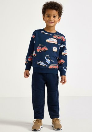 Námořnicky modrá mikina s motivy aut a vrtulníků, doplněná o odpovídající námořnicky modré kalhoty s texturovanými detaily a béžové tenisky.