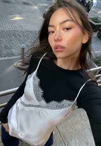 Mladá žena s dlhými tmavými vlasmi, oblečená v čiernom top s dlhým rukávom pod bielym saténovým topom s čipkovým lemovaním, vonku na mestskom chodníku.