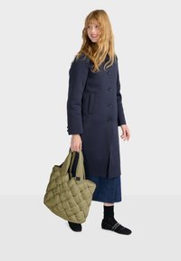 Cappotto di lana navy con bottoni, abbinato a una borsa a tracolla verde trapuntata, gonna di jeans e scarpe slip-on nere, su uno sfondo semplice.