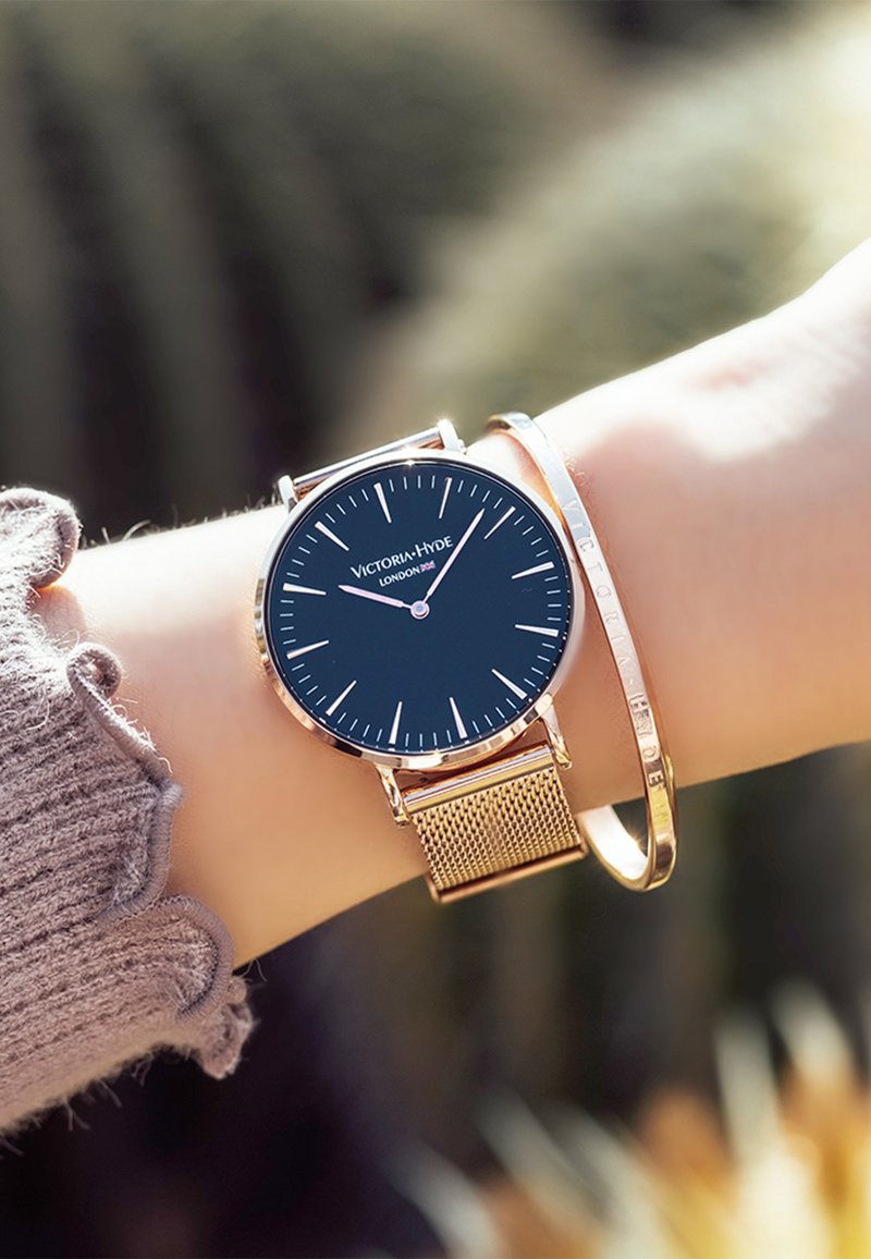 Reloj de esfera negra con correa de malla en oro rosa, con marcadores de hora minimalistas y logotipo. Acompañado de una pulsera delgada en oro.