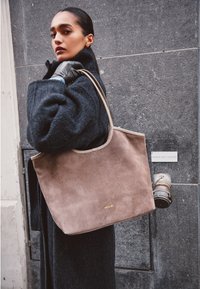 Ruskea mokkanahkainen olkalaukku, jossa on kaareva yläreuna, beige nahkareuna ja kultainen brändäys. Mallilla on päällään musta teksturoitu takki ja hanskat.