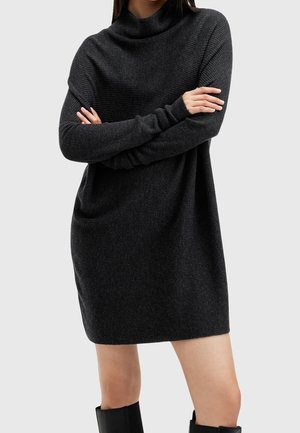 Vrouw draagt een donkergrijze geribbelde gebreide jurk met lange mouwen, armen gekruist en zwarte kniehoge laarzen tegen een effen achtergrond.