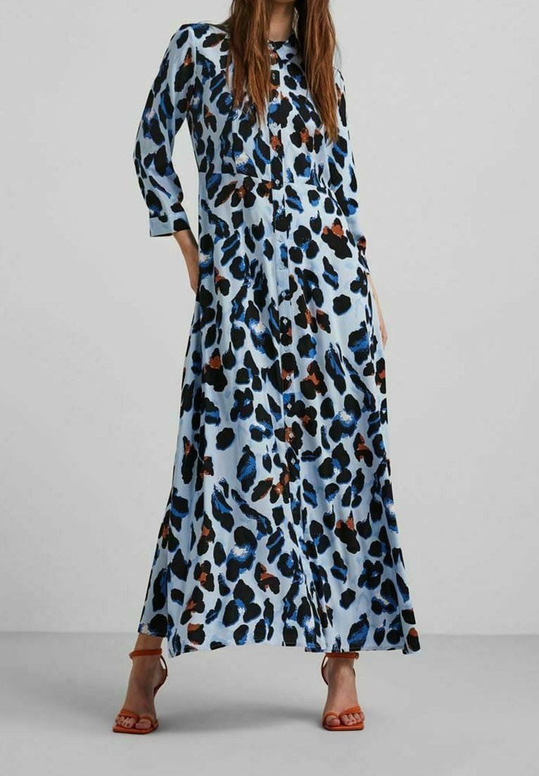 Femme portant une longue robe bleue boutonnée avec des tâches abstraites noires, brunes et bleues, assortie à des sandales à talons hauts orange.