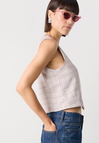 Top cropped fără mâneci de culoare gri deschis cu un model de rețea roz. Materialul este texturat cu un carou subtil, având un decolteu larg și un buzunar pe lateral.