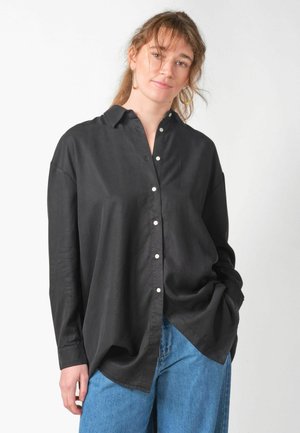 Femeie cu păr castaniu deschis poartă o cămașă neagră largă cu nasturi și blugi albaștri, stând pe un fundal simplu și deschis, având o mână în buzunar.