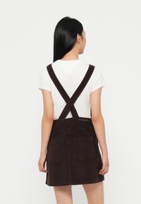 Mørkebrun corduroydress med kryssbelte, to store lommer foran, og "Tommy Jeans" brodert på baksiden. Hvit T-skjorte under.