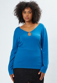 Blauer Langarmpullover mit tiefem V-Ausschnitt und einer Schlitzöffnung, akzentuiert durch ein kreisförmiges Ringdetail am Ausschnitt. Glatte Textur.
