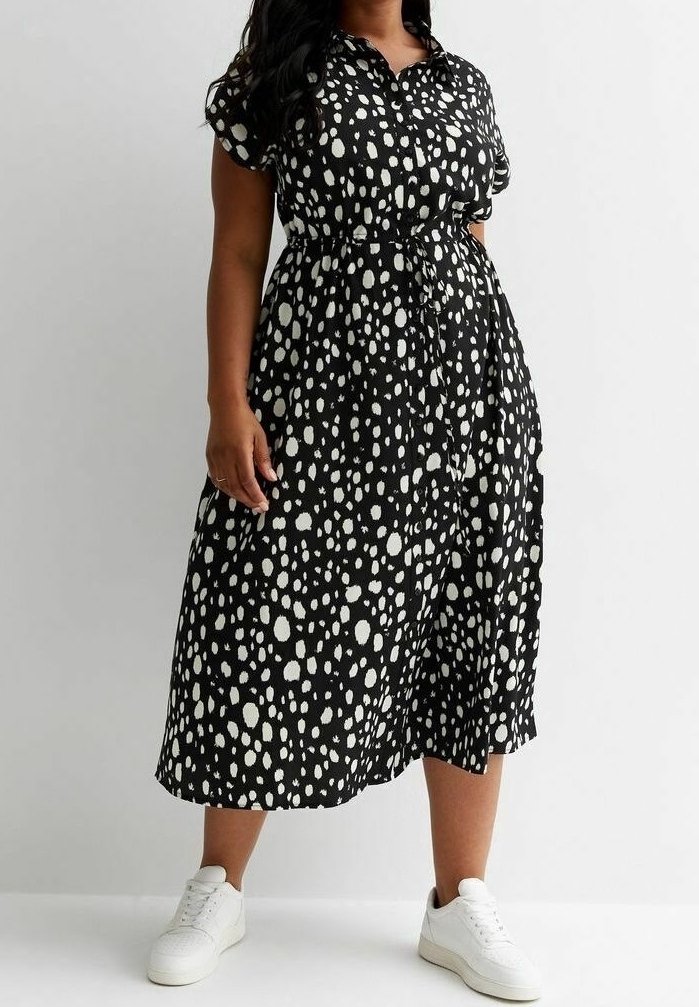 Femme portant une robe midi noire avec des taches blanches irrégulières, manches courtes et baskets blanches, debout devant un fond uni.