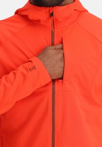 Main en train de fermer la poche poitrine d'une veste zippée orange vif avec capuche et tissu texturé.