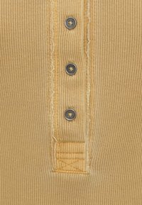 Ύφασμα μουστάρδας με ριμπ υφή, με τέσσερα σκούρα γκρι κουμπιά και ξεφτισμένες άκρες. Διαθέτει ραμμένο μπαλώμα κοντά στην πατιλέτα κουμπιών για επιπλέον λεπτομέρεια.