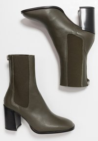 Zielono-oliwkowe botki za kostkę wykonane ze skóry, z elastycznymi panelami bocznymi, spiczastym noskiem i masywnym czarnym obcasem. Teksturowana skórzana faktura.