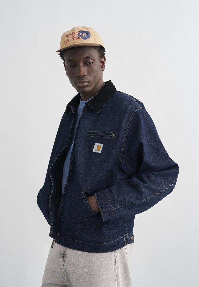 Joven con gorra beige con texto "Heart of Detroit", chaqueta vaquera azul marino con cremallera, camisa azul claro y pantalones gris claro.