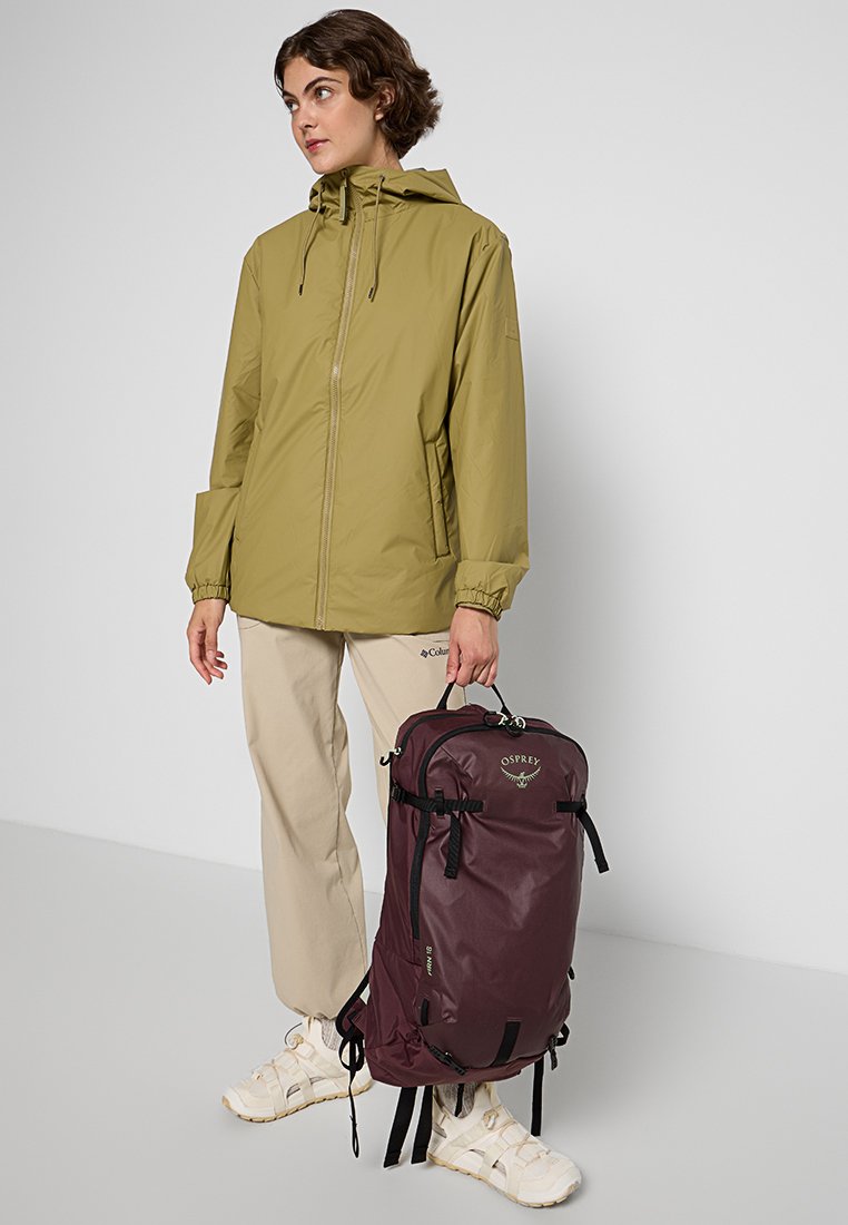 Chaqueta ligera verde oliva con cremallera y puños elásticos; persona sosteniendo una mochila Osprey de color burdeos con correas y cremalleras negras.