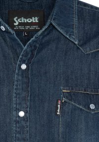 Donkerblauwe denimshirt met een puntige kraag, voorzak, witte knopen en contrasterende gouden stiksels. Bevat een Schott merklabel.