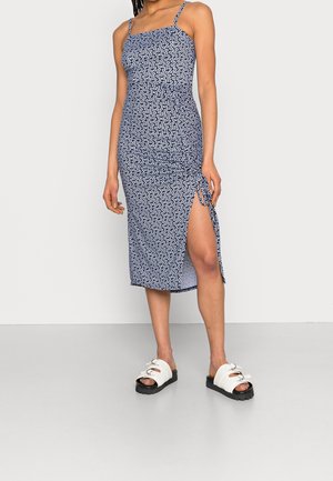 Vrouw met een blauw, bedrukt, mouwloos midijurk met een split aan de zijkant en witte sandalen, staand tegen een egaal lichte achtergrond.