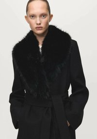 Zwarte jas met een grote nepbonten kraag, taille met ceintuur en lange mouwen. Gestructureerde stof met een gladde afwerking. Minimalistisch ontwerp.