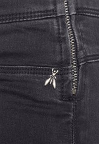 Jeans de mezclilla negros con una cremallera plateada y un alfiler metálico decorativo en la bragueta del bolsillo delantero, con detalles de costuras visibles.