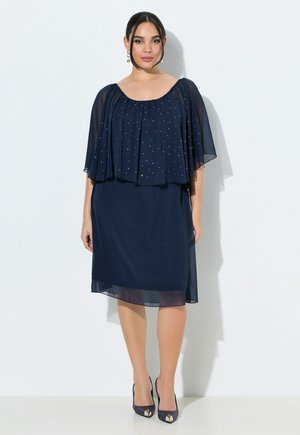 Kobieta w granatowej sukience do kolan z przezroczystym pelerynko ozdobionym małymi srebrnymi kropkami, w parze z granatowymi szpilkami, stojąca na białym tle.